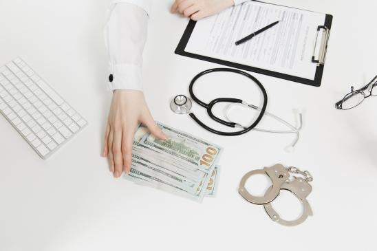 medical fraud, hand reaching for $100 bills alongside handcuffs, stethoscope, clipboard of forms and spectacles 